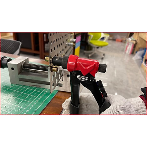 Rote Nietzange beim Nieten an einer Lochplatte. In grauem Handschuh gehalten.